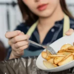 Pessoa com avental despeja batatas fritas de prato branco em lixeira preta, ilustrando desperdício de alimentos em restaurante