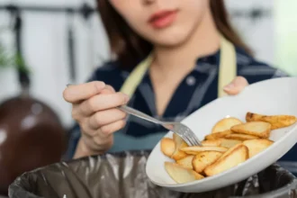 Pessoa com avental despeja batatas fritas de prato branco em lixeira preta, ilustrando desperdício de alimentos em restaurante