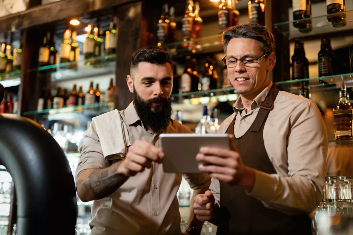 Dois funcionários de avental consultando tablet juntos no balcão de um bar, com prateleiras de garrafas iluminadas ao fundo