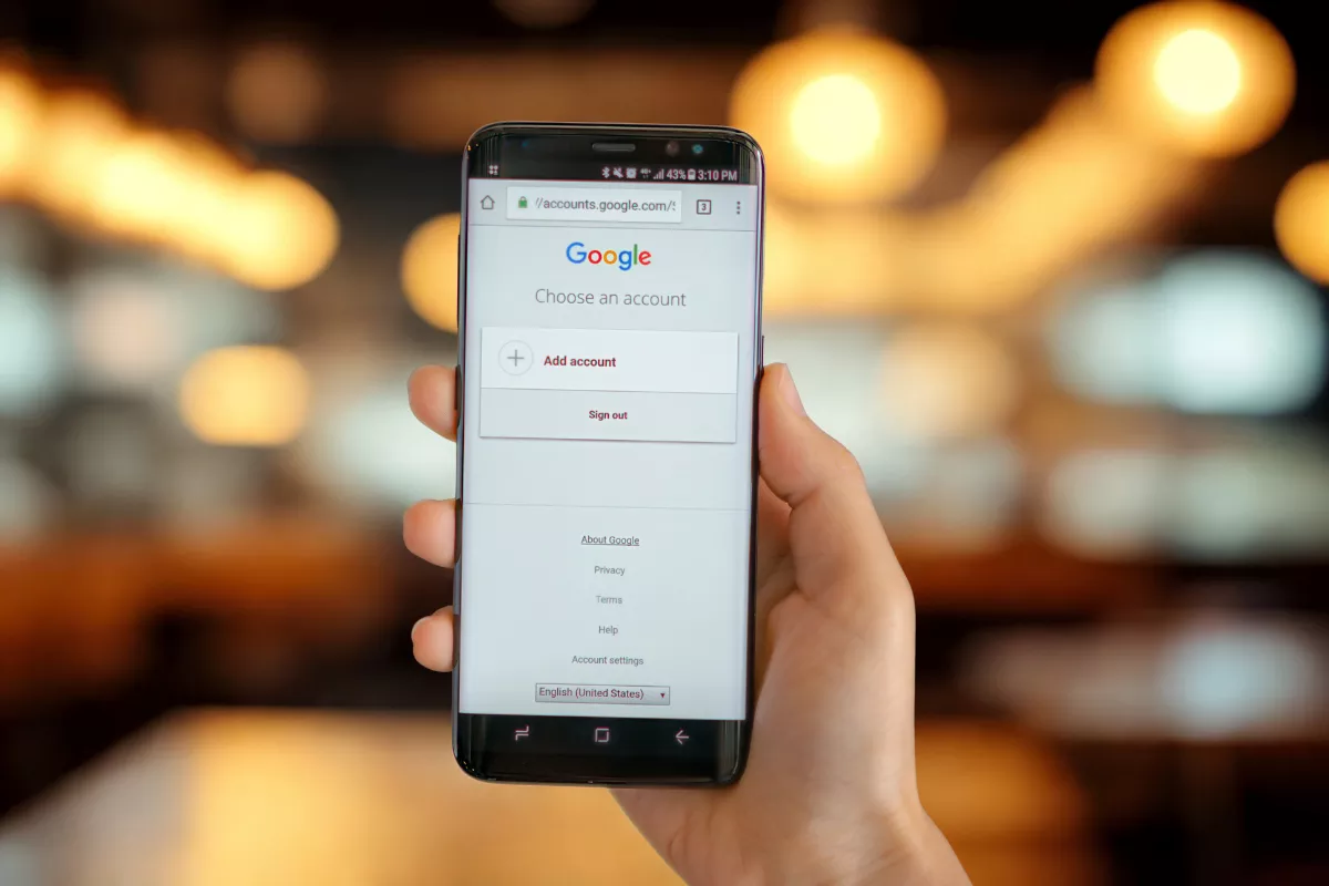 Mão segurando smartphone com tela de login do Google aberta, ambiente de restaurante desfocado ao fundo com luzes quentes