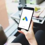 Pessoa sentada segurando smartphone com a tela de abertura do aplicativo Google Ads, mesa com caderno e xícara ao fundo