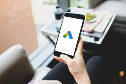 Pessoa sentada segurando smartphone com a tela de abertura do aplicativo Google Ads, mesa com caderno e xícara ao fundo