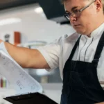 Cozinheiro com avental preto e óculos consultando papéis presos a uma prancheta na cozinha de um restaurante, revisando fichas de produção