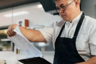Cozinheiro com avental preto e óculos consultando papéis presos a uma prancheta na cozinha de um restaurante, revisando fichas de produção