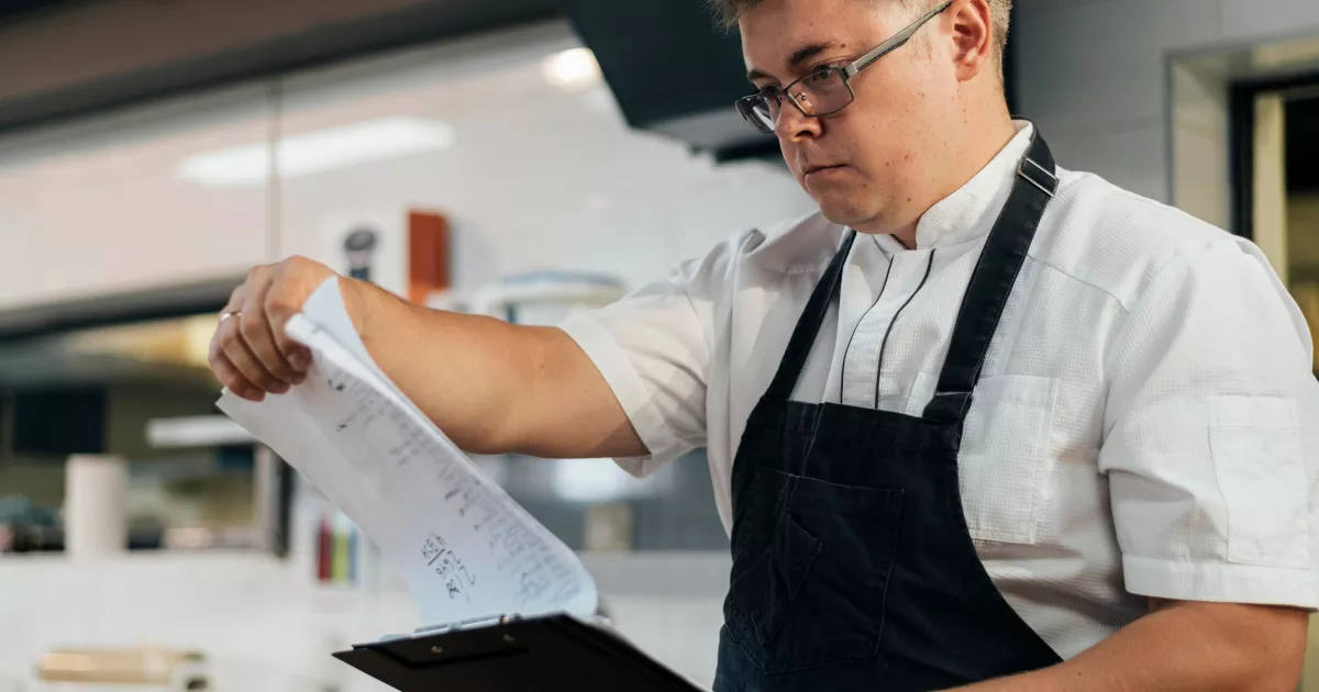 Cozinheiro com avental preto e óculos consultando papéis presos a uma prancheta na cozinha de um restaurante, revisando fichas de produção