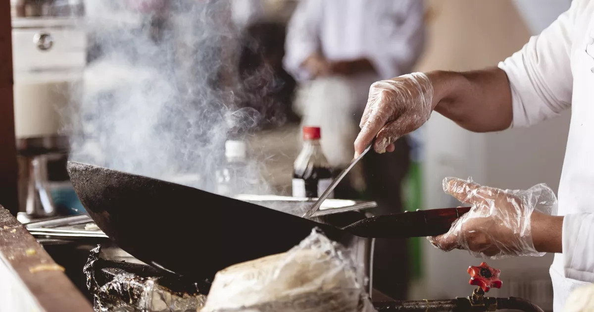 Cozinheiro com luvas descartáveis e jaleco branco mexendo em wok sobre chama alta em fogão industrial, com fumaça subindo e outro profissional ao fundo