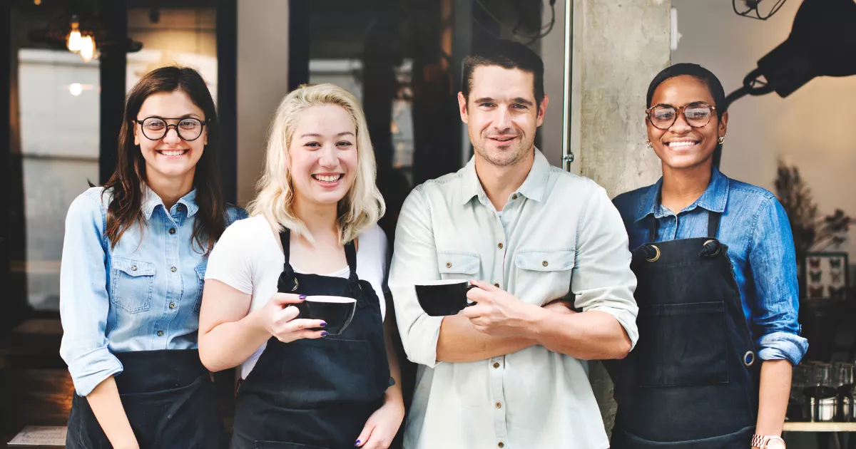 Quatro funcionários de restaurante sorrindo na entrada do estabelecimento, três deles com avental preto, segurando xícaras em ambiente com iluminação quente ao fundo