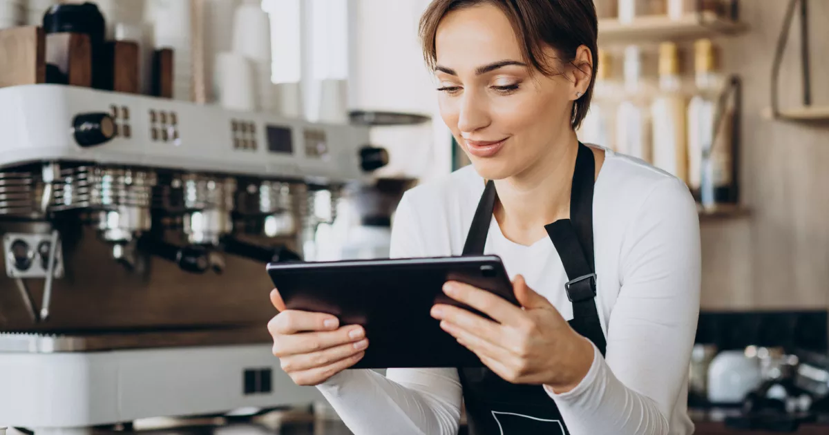 Funcionária de avental preto consultando tablet em frente a máquina de espresso branca, dentro de uma cafeteria com prateleiras ao fundo