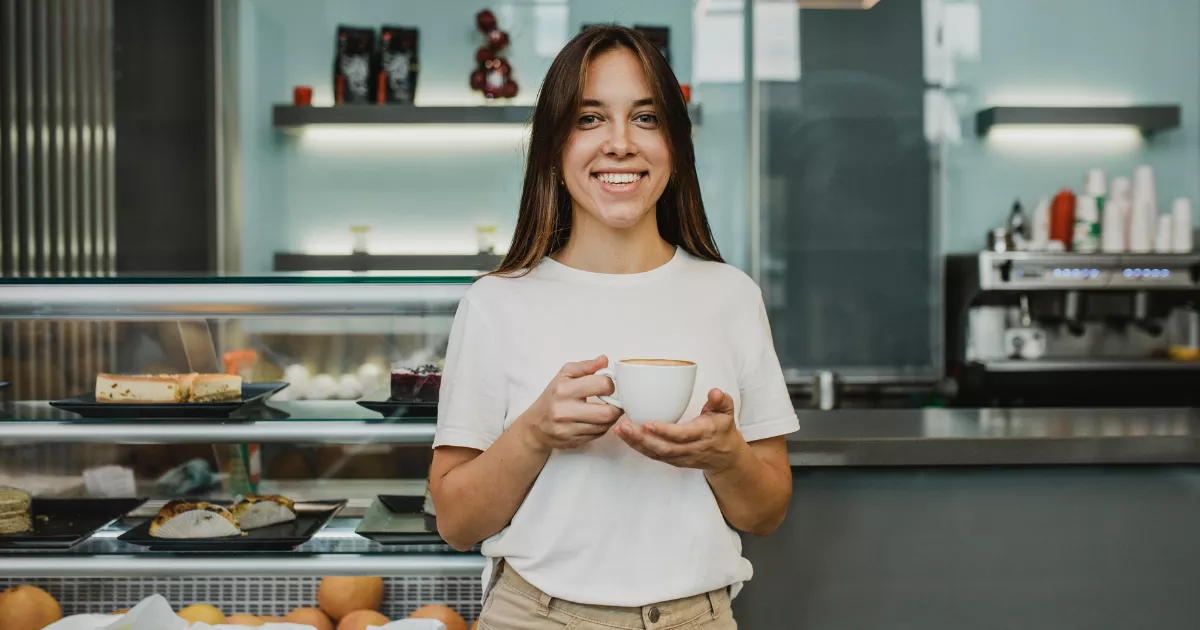 Funcionária de camiseta branca segurando xícara de café com latte art, sorrindo em frente a vitrine com bolos e doces e máquina de espresso ao fundo