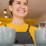 Funcionária de avental cinza e blusa amarela sorrindo atrás do balcão com duas xícaras grandes em primeiro plano, em cafeteria com decoração amarela ao fundo