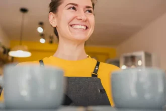 Funcionária de avental cinza e blusa amarela sorrindo atrás do balcão com duas xícaras grandes em primeiro plano, em cafeteria com decoração amarela ao fundo