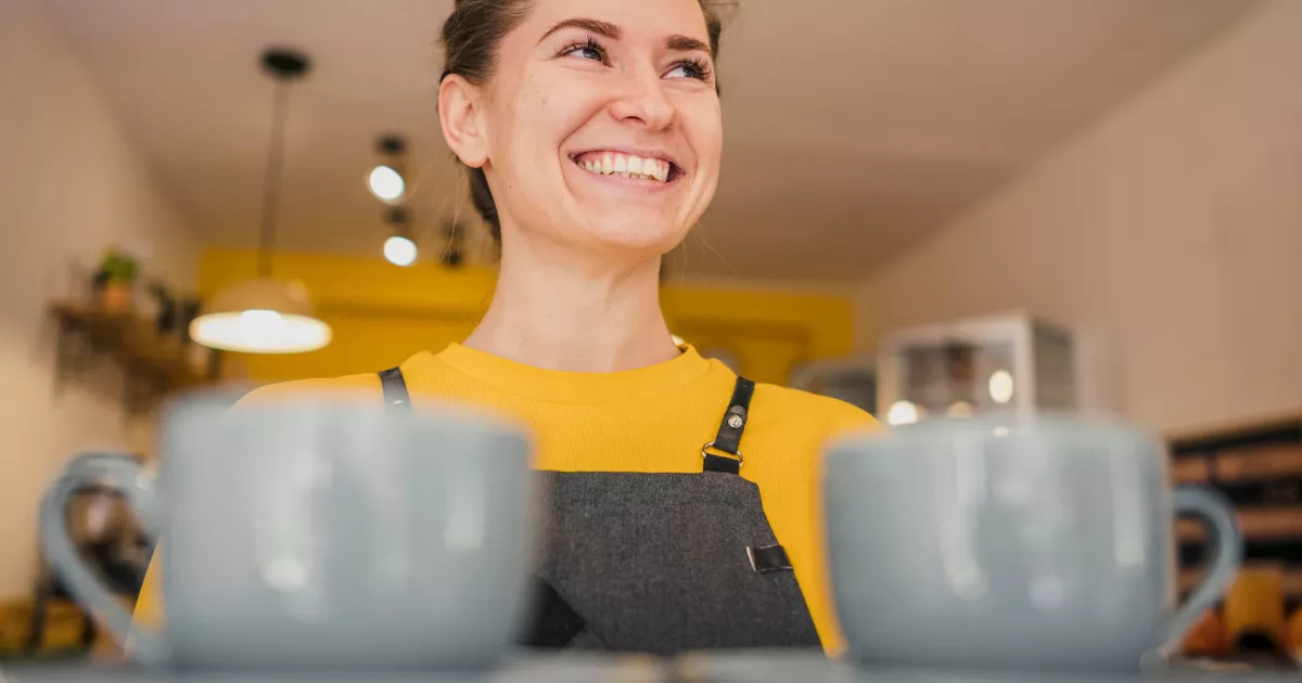 Funcionária de avental cinza e blusa amarela sorrindo atrás do balcão com duas xícaras grandes em primeiro plano, em cafeteria com decoração amarela ao fundo