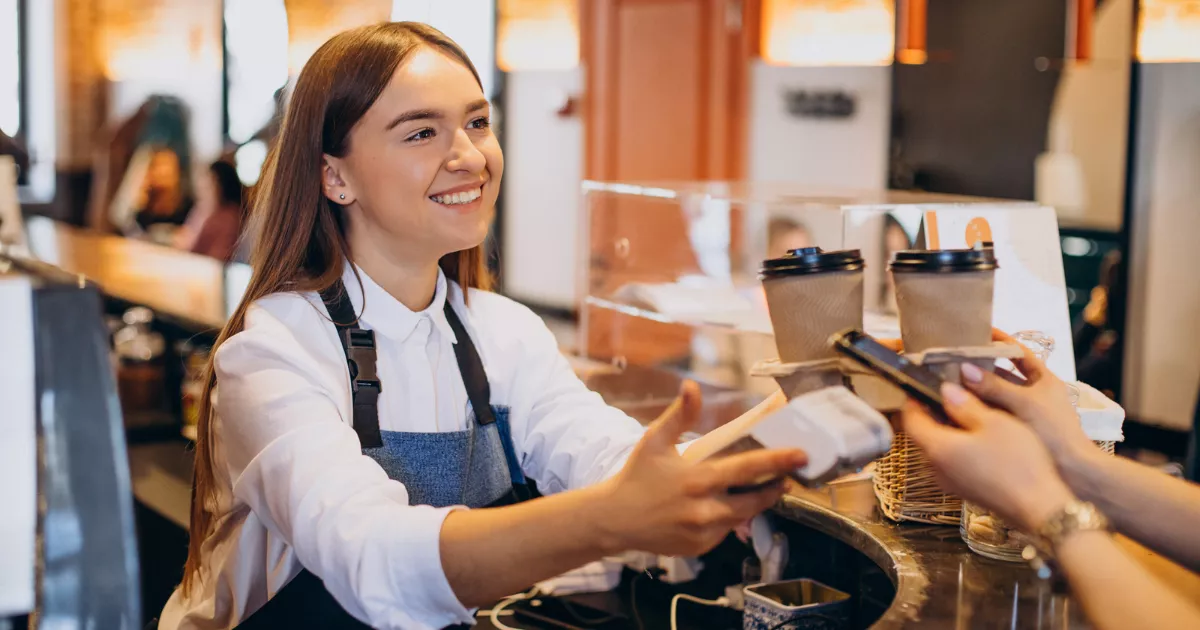 Atendente de cafeteria com avental azul entregando maquininha de cartão ao cliente no balcão, com copos descartáveis e itens de confeitaria ao fundo