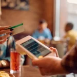 Cliente segurando cartão próximo a maquininha de pagamento apresentada por garçom em mesa de restaurante, com família ao fundo em ambiente iluminado