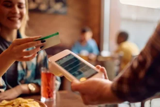 Cliente segurando cartão próximo a maquininha de pagamento apresentada por garçom em mesa de restaurante, com família ao fundo em ambiente iluminado