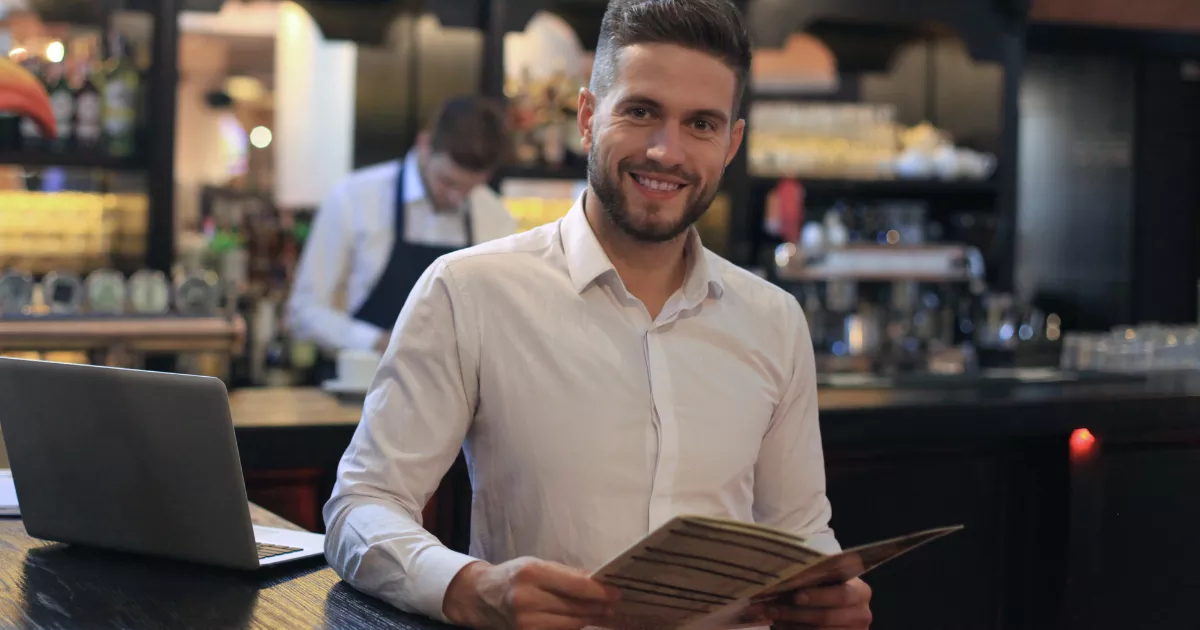 Dono de restaurante de camisa branca segurando cardápio e sorrindo ao balcão, com notebook aberto ao lado e funcionário de avental trabalhando ao fundo