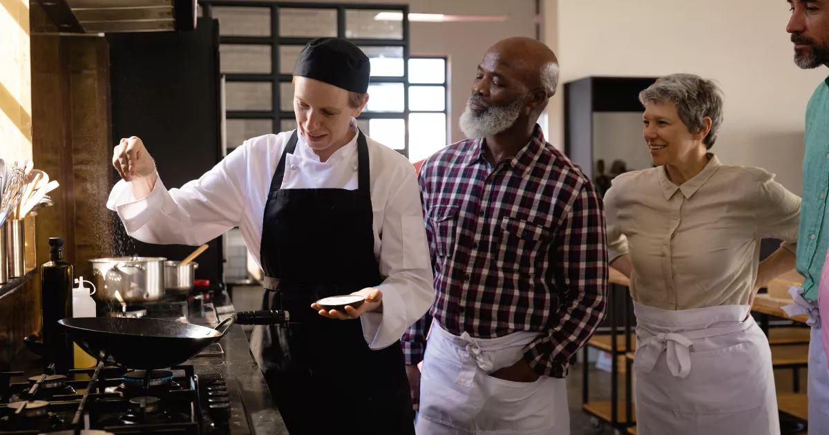 Chef de jaleco branco e avental preto temperando prato ao fogão enquanto três funcionários de avental observam a demonstração na cozinha do restaurante