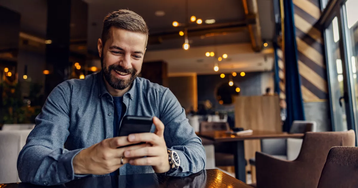 Homem de barba sorri ao olhar o celular sentado em mesa de restaurante com iluminação aconchegante e luminárias pendentes ao fundo, usando o ChatGPT no dia a dia do negócio.