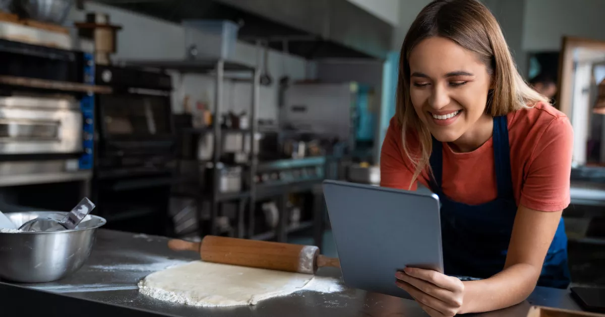 Cozinheira sorri olhando o tablet enquanto abre massa de pizza com rolo na bancada da cozinha, gerenciando pedidos e conteúdo do restaurante entre as tarefas