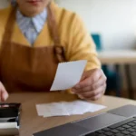 Gestora de restaurante com avental conferindo cupom fiscal ao lado de celular e notebook na mesa, organizando o fechamento fiscal do período