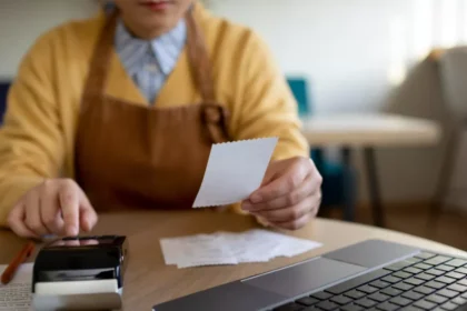 Gestora de restaurante com avental conferindo cupom fiscal ao lado de celular e notebook na mesa, organizando o fechamento fiscal do período