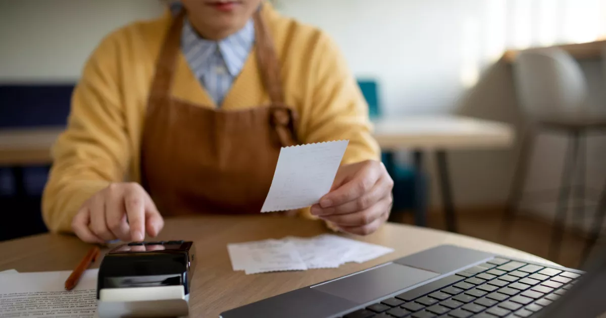 Gestora de restaurante com avental conferindo cupom fiscal ao lado de celular e notebook na mesa, organizando o fechamento fiscal do período
