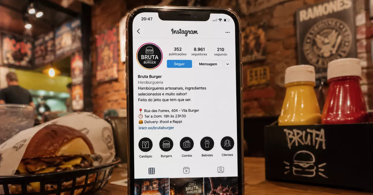 Celular mostra a bio comercial da Bruta Burger no Instagram, com descrição, endereço, horário, delivery e link de pedidos, ao lado de molhos e hambúrguer na hamburgueria