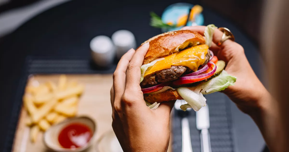 Mãos seguram hambúrguer artesanal com queijo derretido, cebola roxa, tomate e alface, com batata frita e ketchup ao fundo, exemplo de foto de prato apetitosa para o Instagram