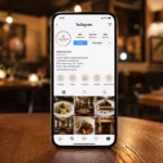 Celular exibe o perfil comercial do restaurante Sabores da Casa no Instagram, com bio, endereço, horário e Destaques de cardápio, sobre mesa de madeira no salão iluminado