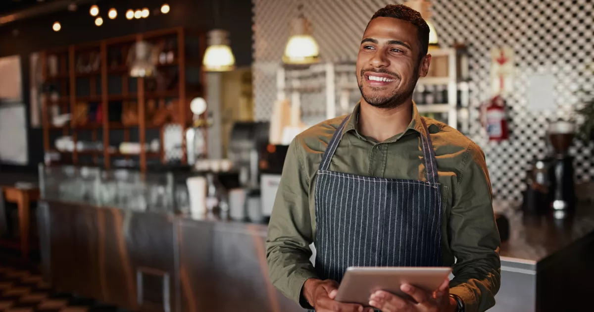Dono de restaurante com avental listrado segurando tablet, sorrindo, de pé em frente ao balcão com banquetas e prateleiras ao fundo
