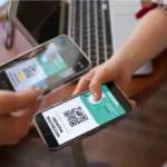 Duas pessoas trocando celulares enquanto uma escaneia o QR code exibido na tela da outra, notebook e plantas ao fundo sobre mesa de madeira
