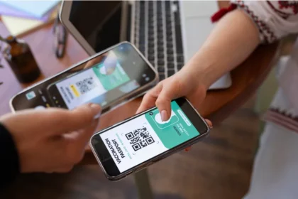 Duas pessoas trocando celulares enquanto uma escaneia o QR code exibido na tela da outra, notebook e plantas ao fundo sobre mesa de madeira