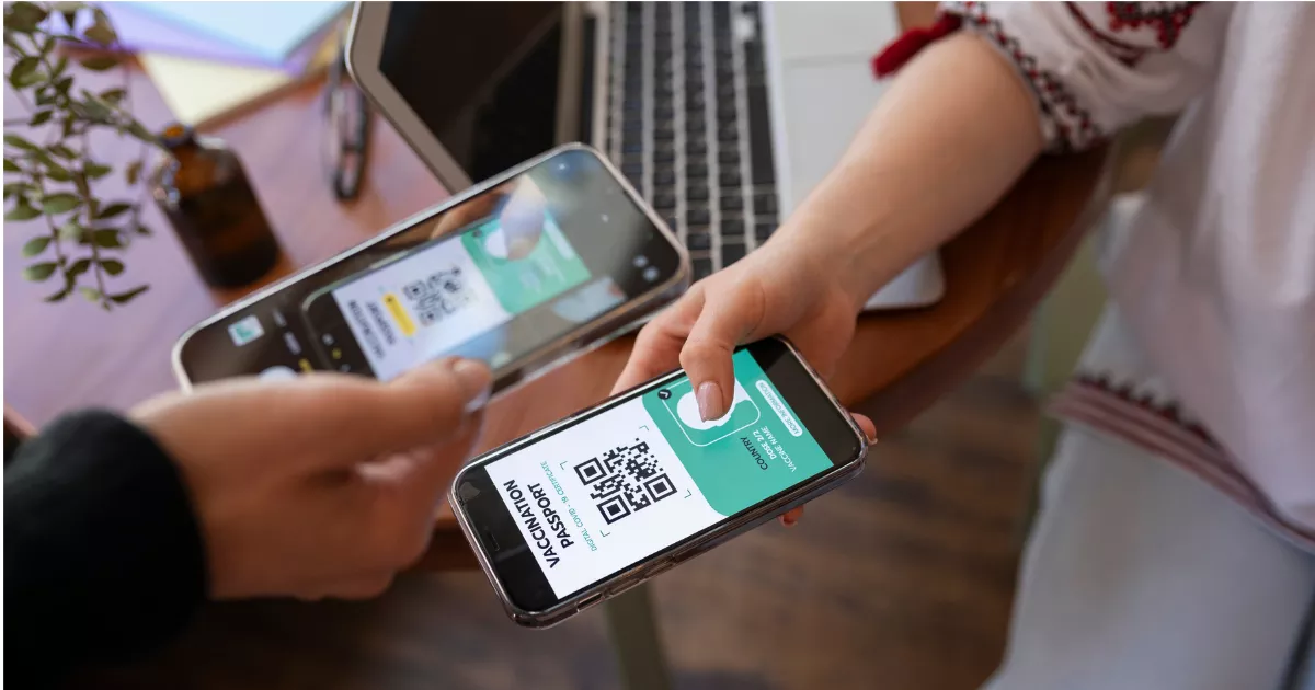 Duas pessoas trocando celulares enquanto uma escaneia o QR code exibido na tela da outra, notebook e plantas ao fundo sobre mesa de madeira