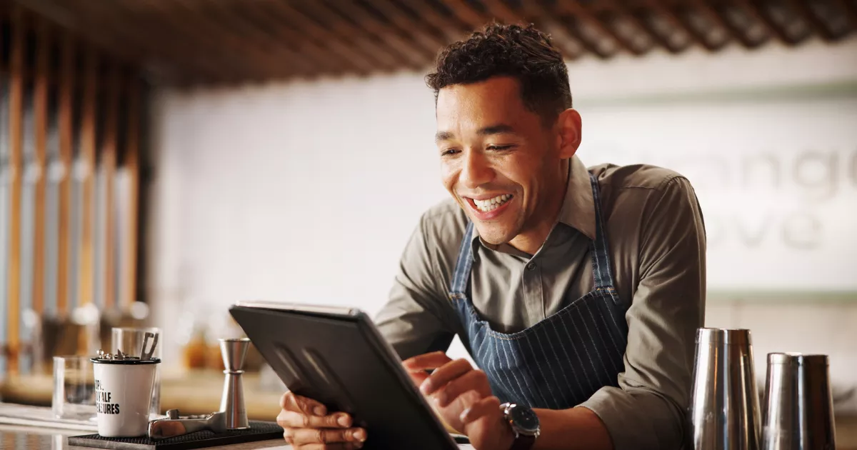 Atendente de avental listrado sorrindo enquanto consulta tablet apoiado no balcão de um bar, com coqueteleiras e utensílios ao lado, conferindo pedidos de delivery