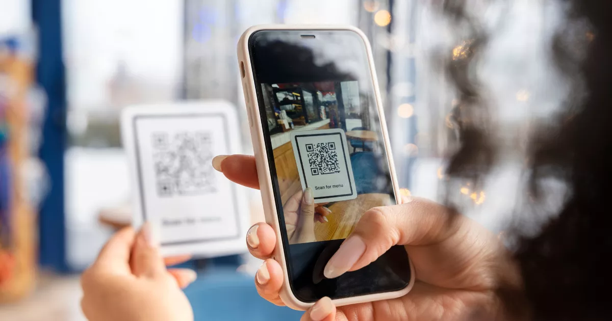 Mulher de cabelos cacheados usando celular para escanear QR code em suporte de mesa, interior de café com luzes e decoração de madeira ao fundo