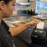 Atendente de avental preto retira o cupom recém-impresso de uma impressora térmica Bematech no balcão de uma lanchonete, com computador do caixa ao lado