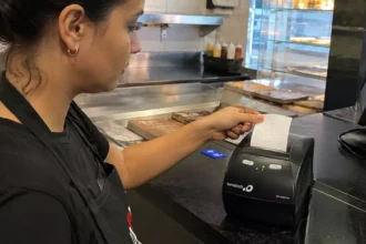 Atendente de avental preto retira o cupom recém-impresso de uma impressora térmica Bematech no balcão de uma lanchonete, com computador do caixa ao lado