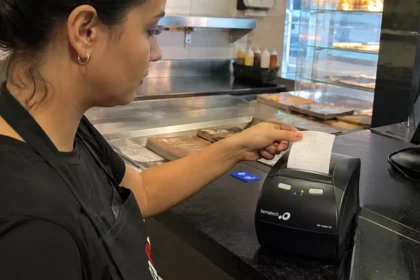 Atendente de avental preto retira o cupom recém-impresso de uma impressora térmica Bematech no balcão de uma lanchonete, com computador do caixa ao lado