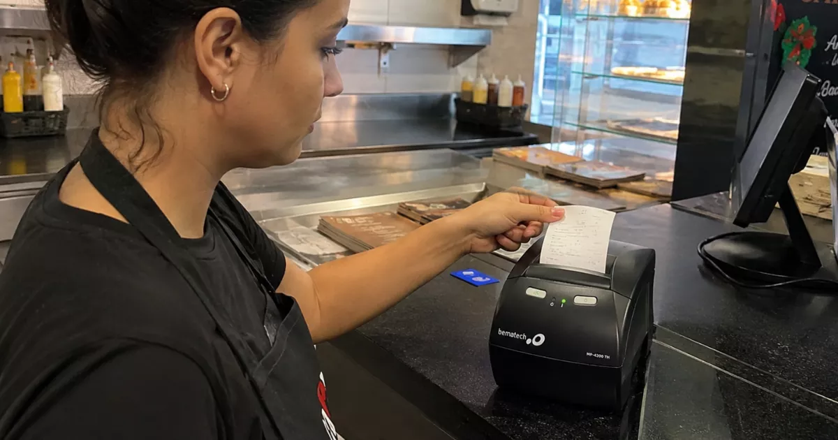 Atendente de avental preto retira o cupom recém-impresso de uma impressora térmica Bematech no balcão de uma lanchonete, com computador do caixa ao lado