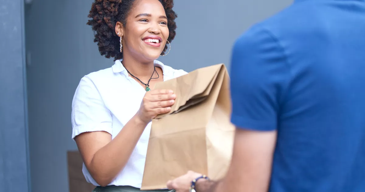 Cliente sorrindo ao receber grande sacola kraft de um entregador na porta de casa, com a mão do entregador parcialmente visível em primeiro plano