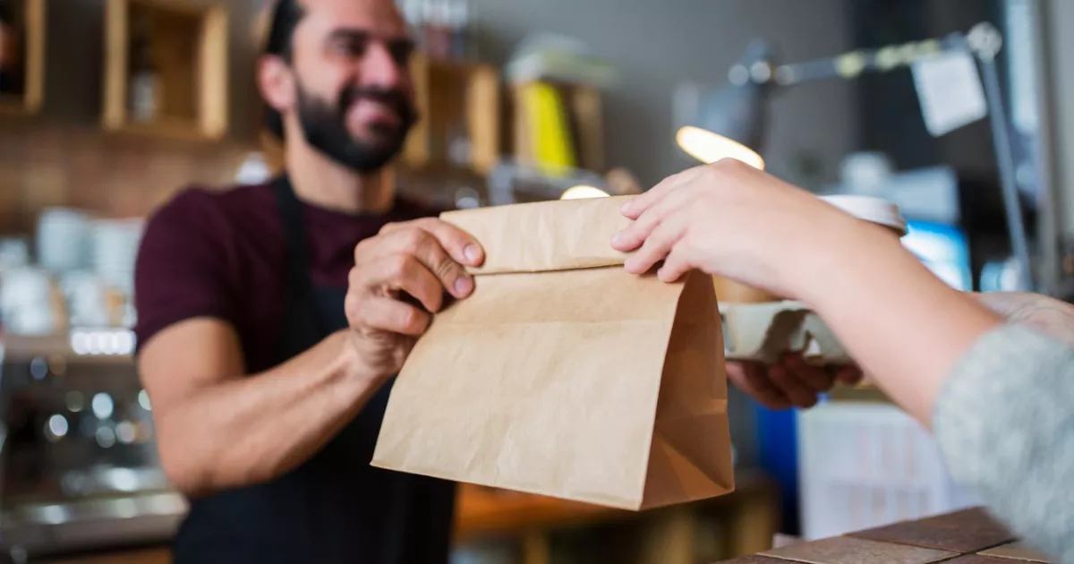 Funcionário de restaurante com avental escuro entregando sacola kraft no balcão para cliente que também recebe copo de café, com o interior do estabelecimento desfocado ao fundo