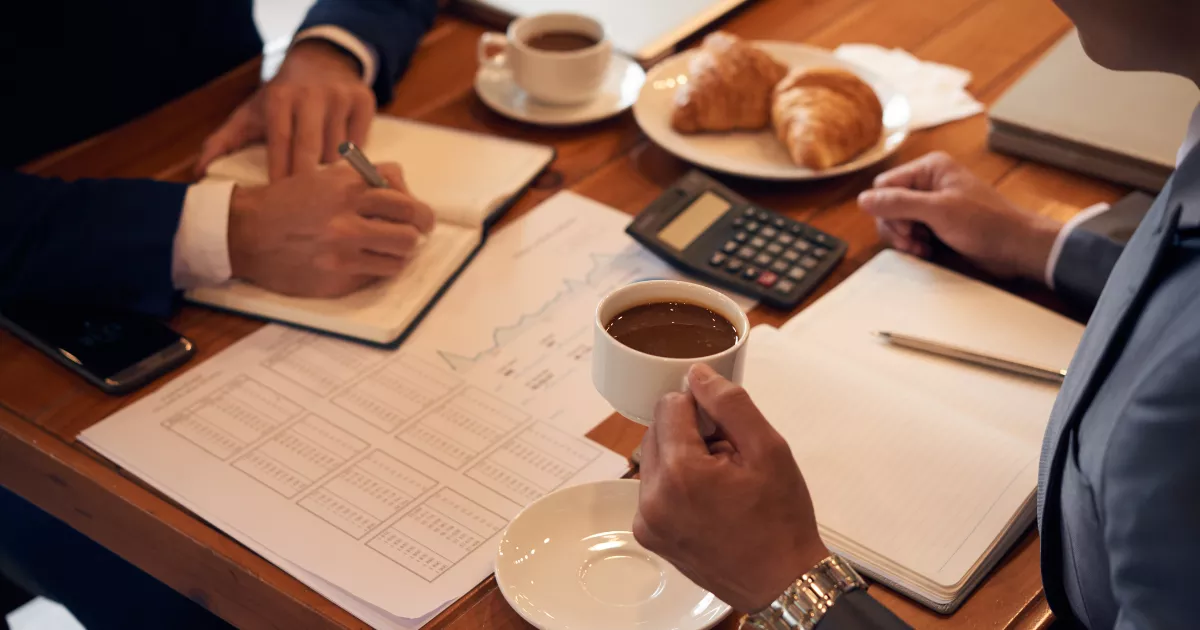 Duas pessoas de terno analisam planilhas financeiras com gráficos sobre mesa de madeira, com calculadora, cadernos, café e croissants, em reunião de gestão financeira de restaurante.