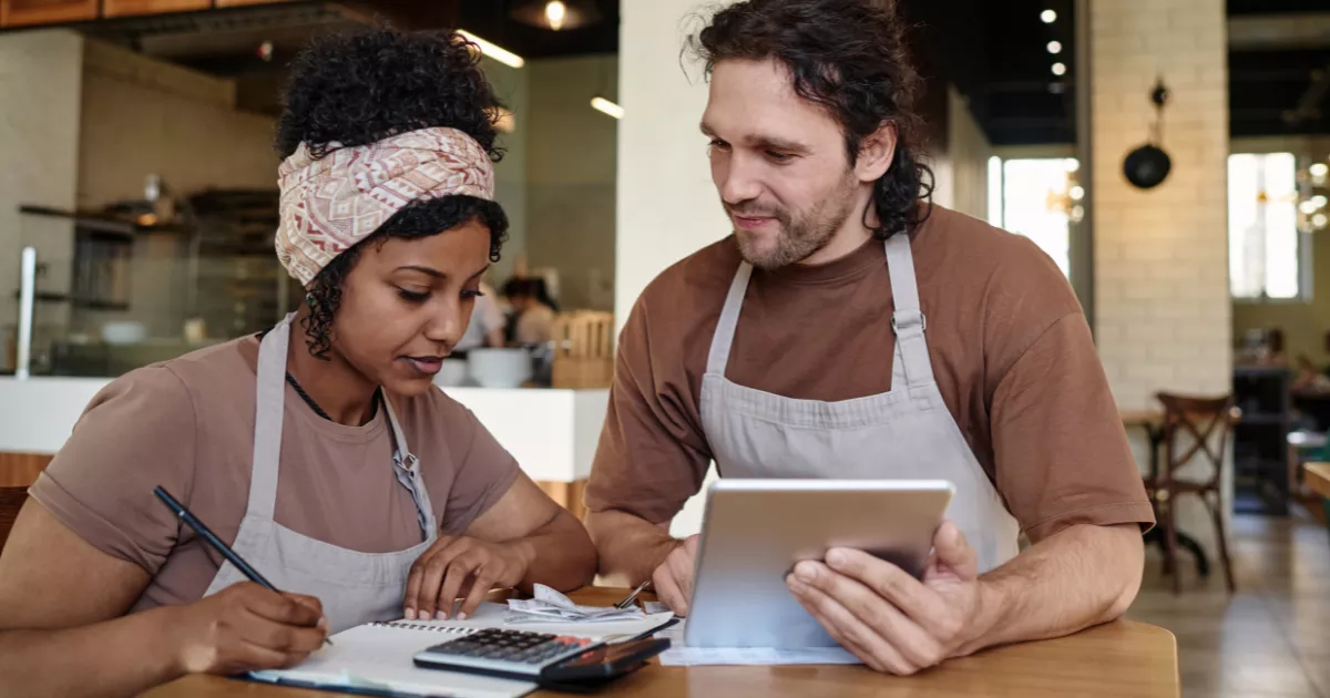 Dois funcionários com avental consultam tablet e fazem anotações sentados à mesa, com calculadora sobre o caderno e cozinha aberta desfocada ao fundo, revisando custos do restaurante.
