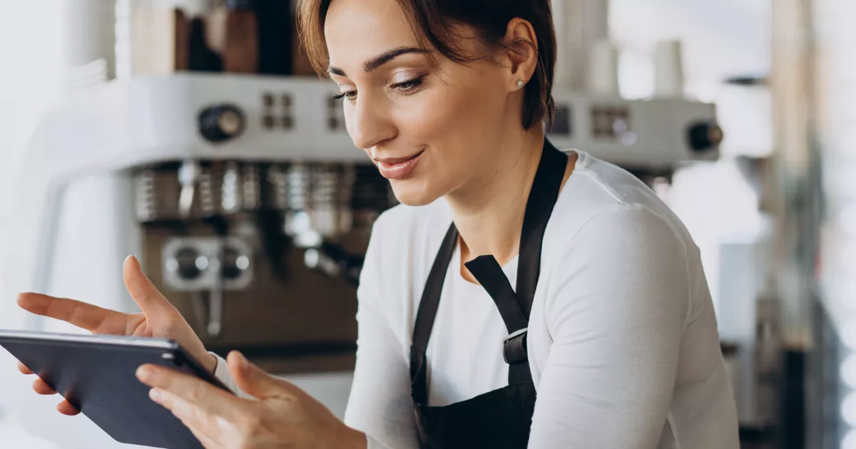 Mulher com avental preto sorri ao consultar tablet atrás do balcão de café, com máquina de espresso desfocada ao fundo, acompanhando os números do estabelecimento.