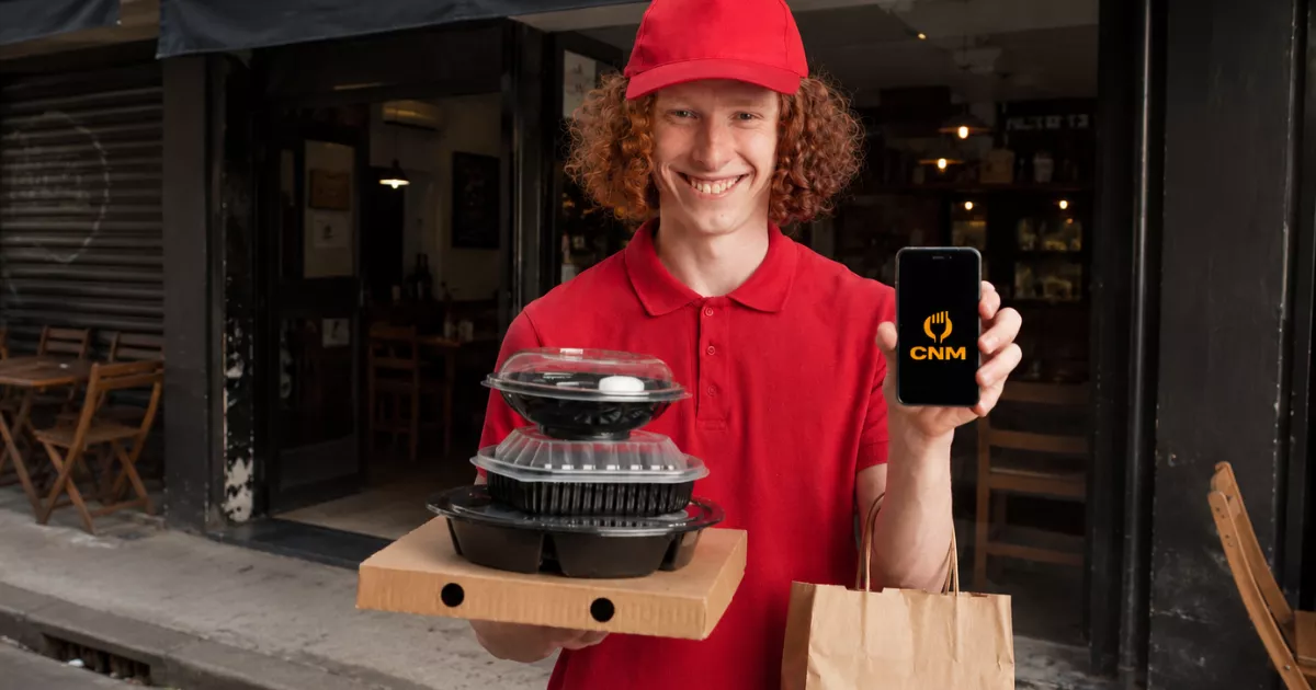 Entregador de uniforme vermelho segurando embalagens de delivery empilhadas e mostrando o celular com o aplicativo ControleNaMão aberto, na porta do restaurante