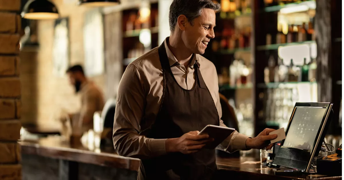 Gestor de restaurante de avental confere pedidos no tablet enquanto opera o terminal do caixa, com balcão do bar e prateleiras de bebidas ao fundo
