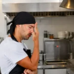 Cozinheiro de bandana preta e avental com a mão no rosto, expressão de cansaço, parado na cozinha profissional durante o horário de pico do restaurante