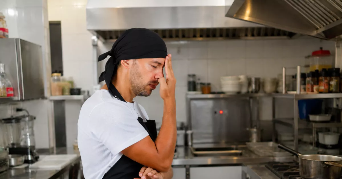 Cozinheiro de bandana preta e avental com a mão no rosto, expressão de cansaço, parado na cozinha profissional durante o horário de pico do restaurante