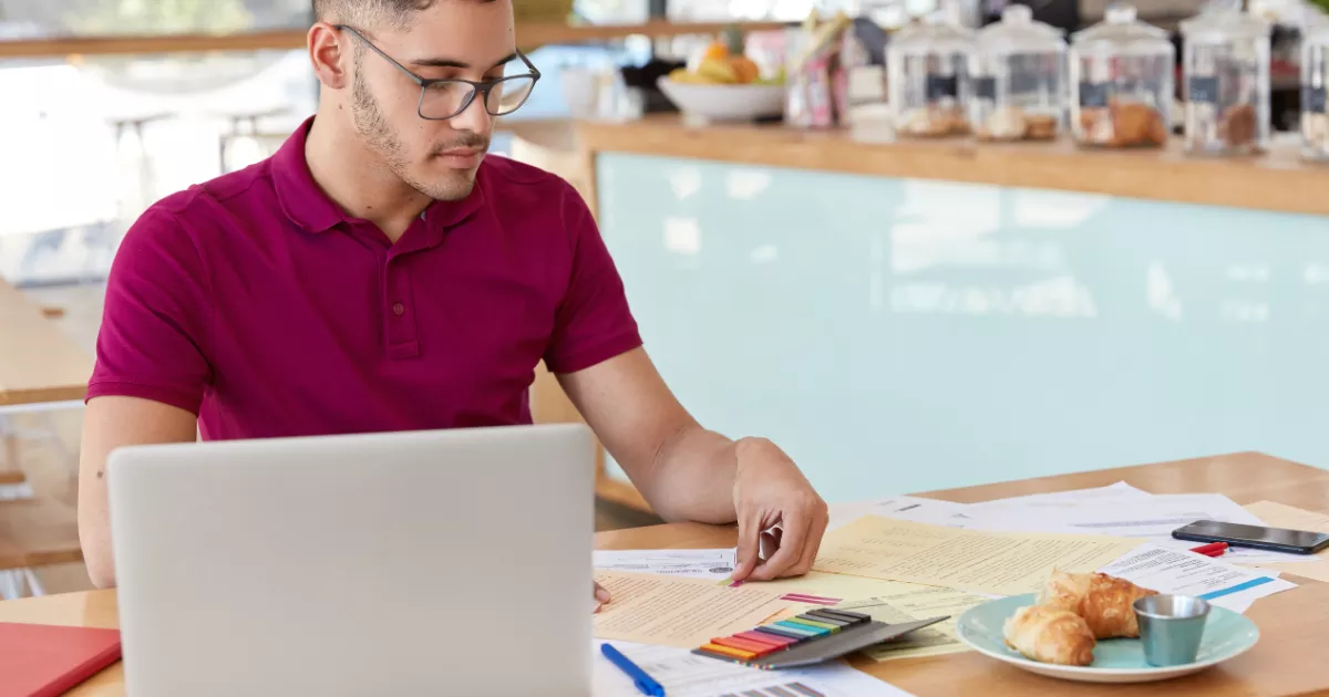 Jovem de óculos e camisa polo vinho revisa documentos financeiros impressos com canetas coloridas, com notebook aberto e balcão de café com potes ao fundo.