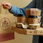 Pessoa de jaqueta jeans segurando conjunto de embalagens personalizadas de confeitaria com logotipo impresso em sacola kraft, potes e caixa prontos para entrega de delivery